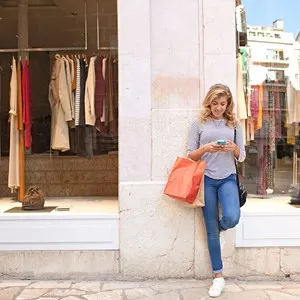 Beautiful teenager young woman in city shopping street by fashion store window, smiling using a smartphone to network online, outdoors. Adolescent consumer using technology, lifestyle exterior.