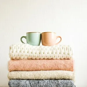 Two cups of coffee standing on bunch of knitted warm pastel color sweaters