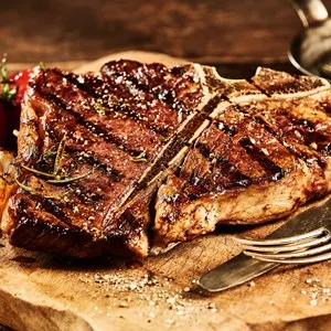 Succulent grilled large t-bone steak garnished with herbs, tomato and salt with fork and knife beside it on cutting board