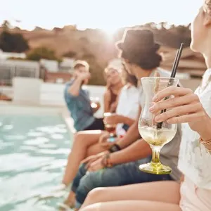 Woman holding a cocktail glass while sitting on the edge of swimming pool with friends. Young people enjoying a poolside party with drinks. Focus on hand and cocktail glass.