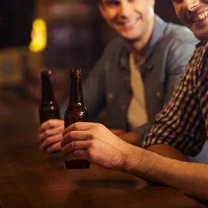 Two young men in casual clothes are smiling, looking at camera and holding bottles of beer while sitting at bar counter in pub, close-up